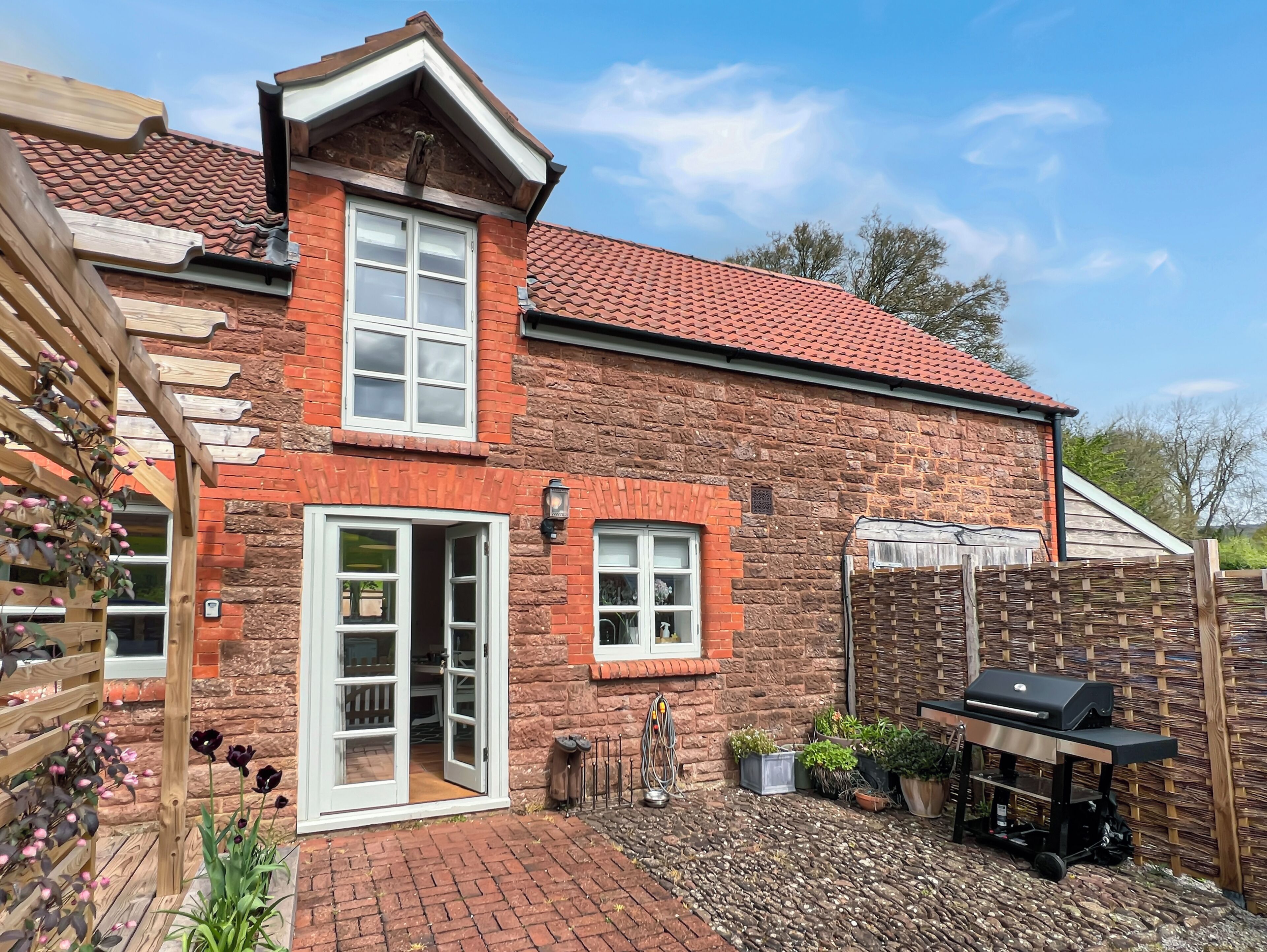 A brick house with a red-tiled roof, a wooden pergola with flowers, a barbecue grill, and a stone wall.