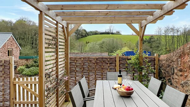 Een houten terras met een tafel, stoelen, een fles wijn en een schaal fruit.
