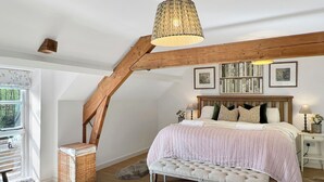 Een slaapkamer met een schuine plafond, een bed met houten hoofdeinde, een bankje, een rieten mand en een raam met zonwering.