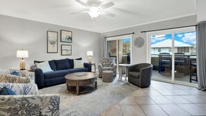 Un salon spacieux doté d’un ventilateur de plafond, d’une grande fenêtre offrant une vue sur l’extérieur et d’un coin repas avec des chaises et une table.