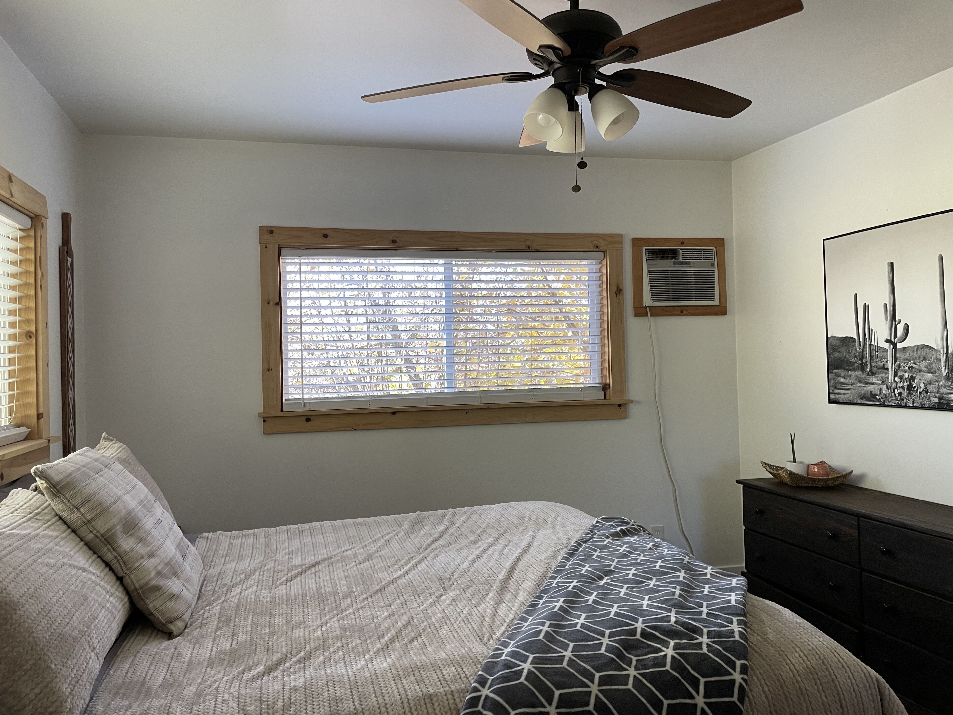 Slaapkamer met een bed, een plafondventilator, een raam met jaloezieën, een dressoir en een wandairconditioner.