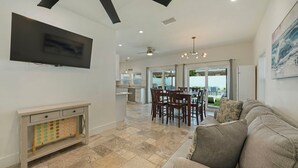 A modern living room with a flat-screen TV, a fireplace, a dining area with chairs, and a large window offering a view of the backyard. - A Wave From It All - Beach Cottage with Private Pool & Outdoor Area (Miramar Beach)