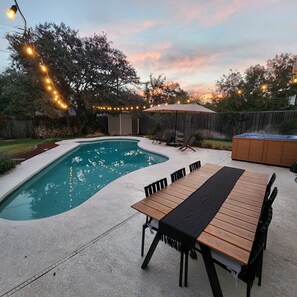 Un patio trasero con piscina, una mesa de madera y sillas. Hay luces colgantes y una sombrilla.