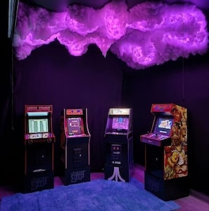 Cuatro máquinas de arcade en una habitación con iluminación morada y un adorno en el techo que parece una nube.