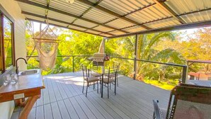 Área externa coberta com mesa e cadeiras, luminária pendente e vista para o verde.