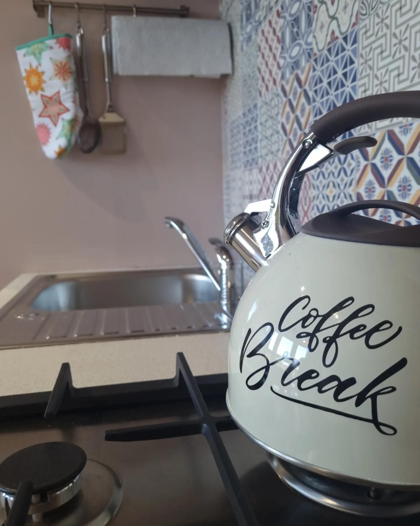 A kitchen with a kettle labeled "Coffee Break" on a stove, a sink, and a colorful mug hanging on the wall.
