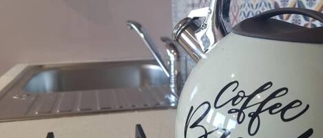 A kitchen with a kettle labeled "Coffee Break" on a stove, a sink, and a colorful mug hanging on the wall.