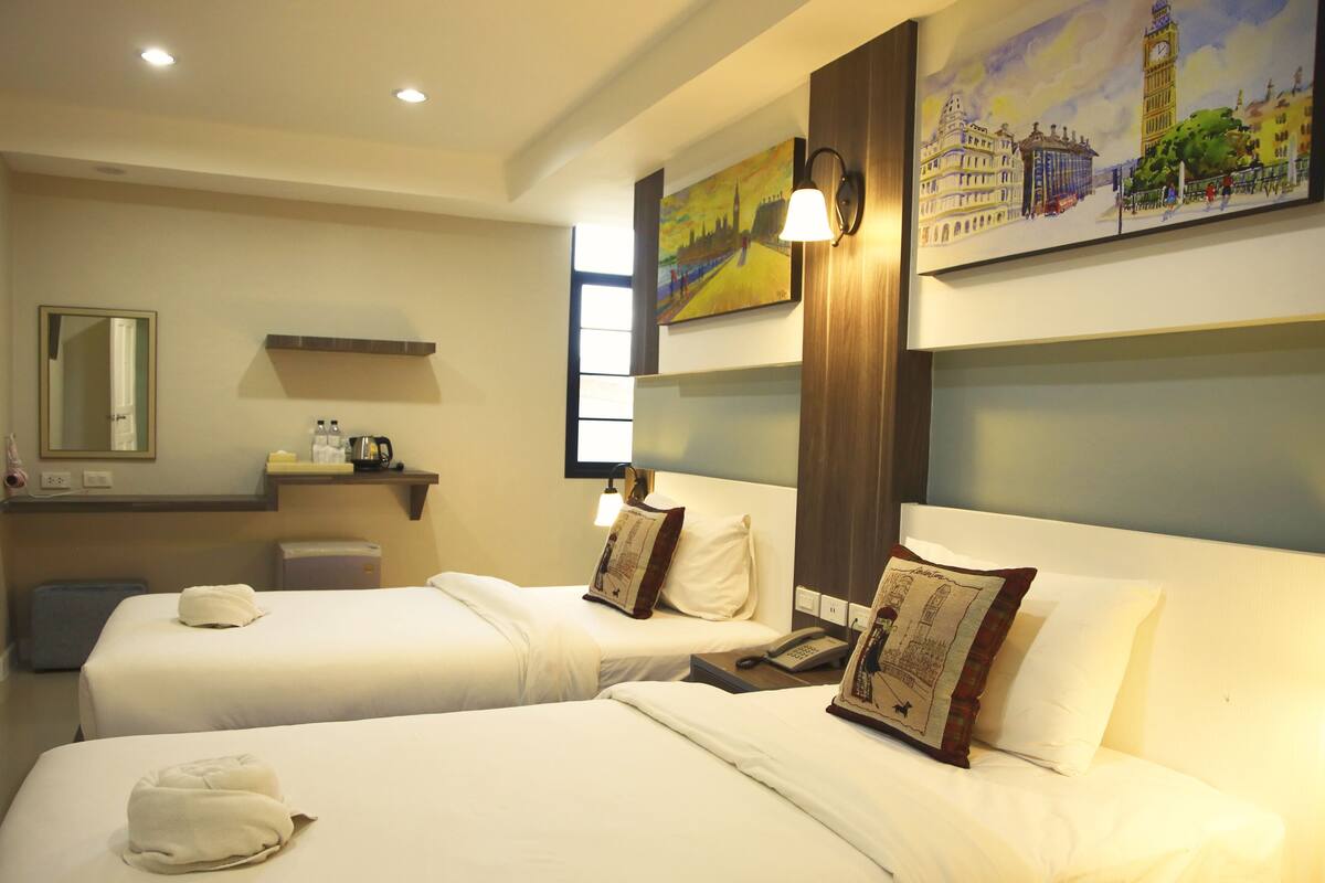 A hotel room with two beds, a bedside table, a lamp, a painting, and a mirror.