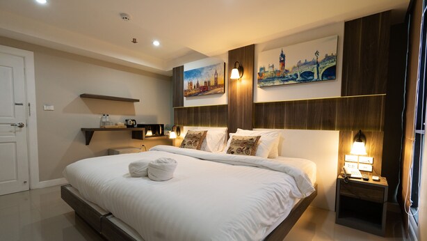 Una habitación de hotel moderna con una cama grande, una mesita de noche con lámpara, un estante en la pared y dos cuadros de paisajes urbanos.