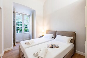 A hotel room with two beds, a nightstand, a lamp, and a door leading to a balcony with a view of greenery.