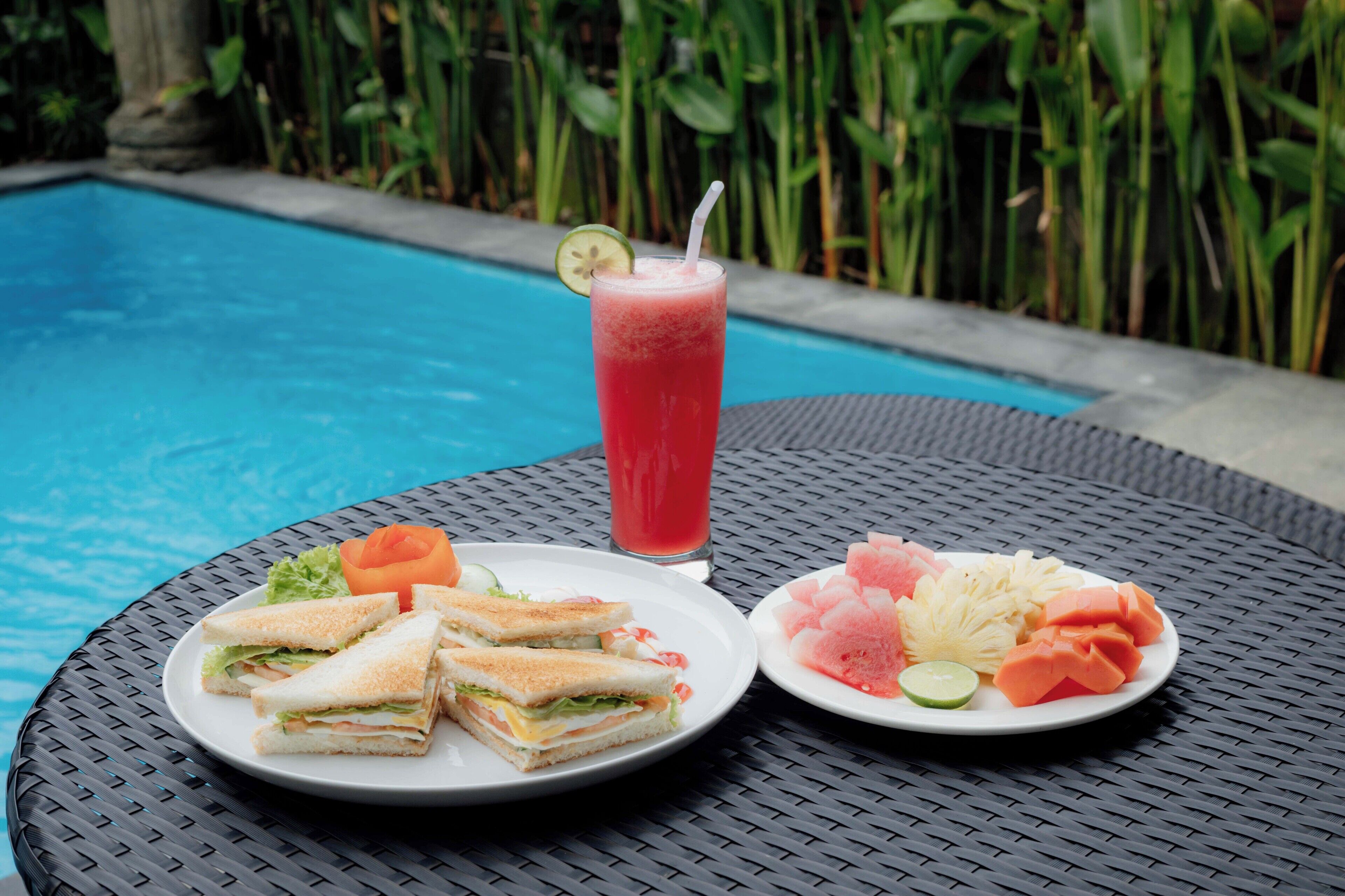 Une table avec une assiette de sandwichs, une assiette de fruits et un verre d’une boisson rouge, le tout placé près d’une piscine.