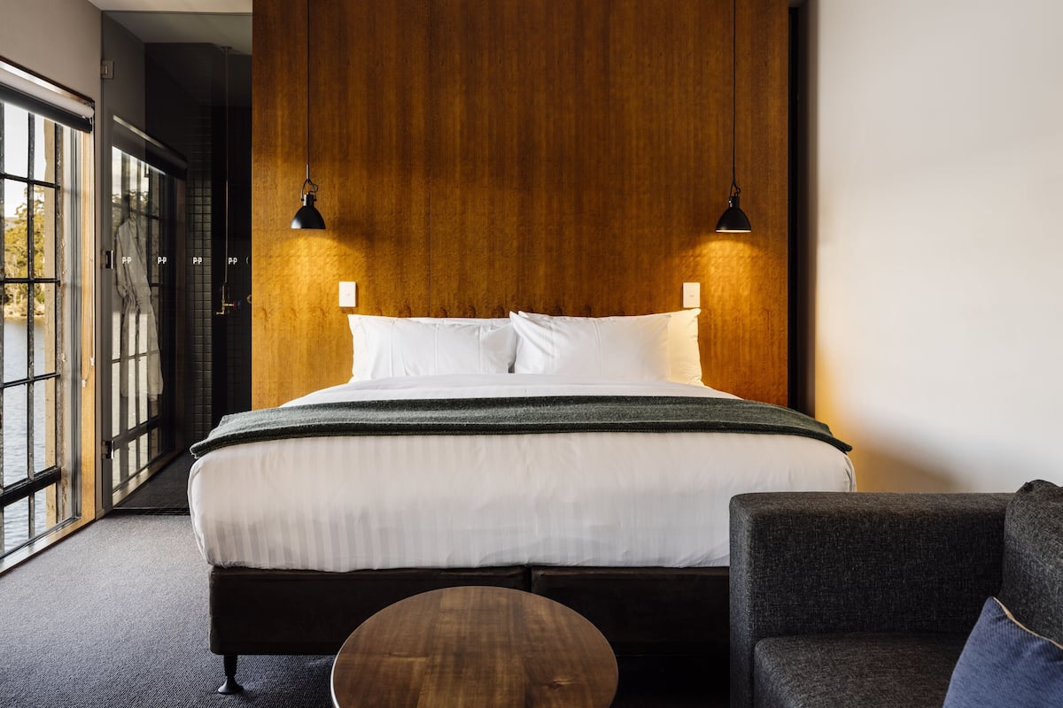 Habitación de hotel moderna con una cama grande, dos lámparas colgantes, un sofá oscuro y vistas a través de un ventanal.
