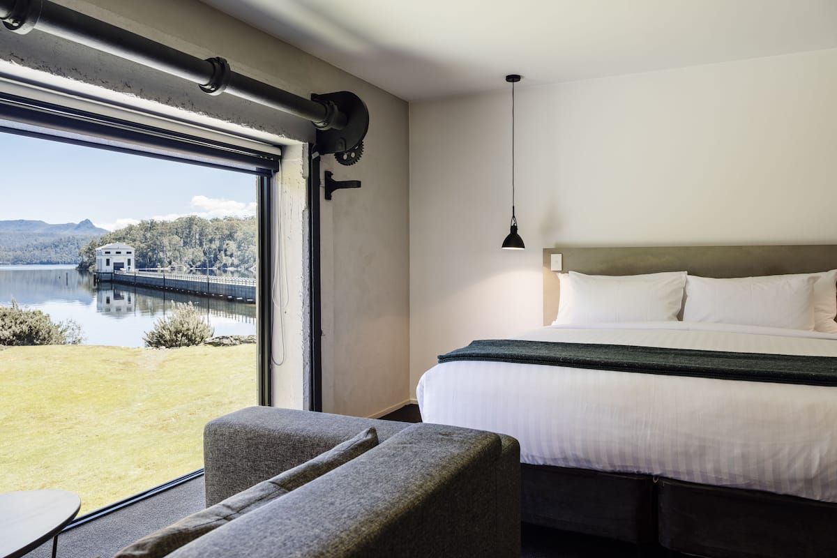 Habitación de hotel con un ventanal grande, una cama, un escritorio y vistas a un lago y montañas.