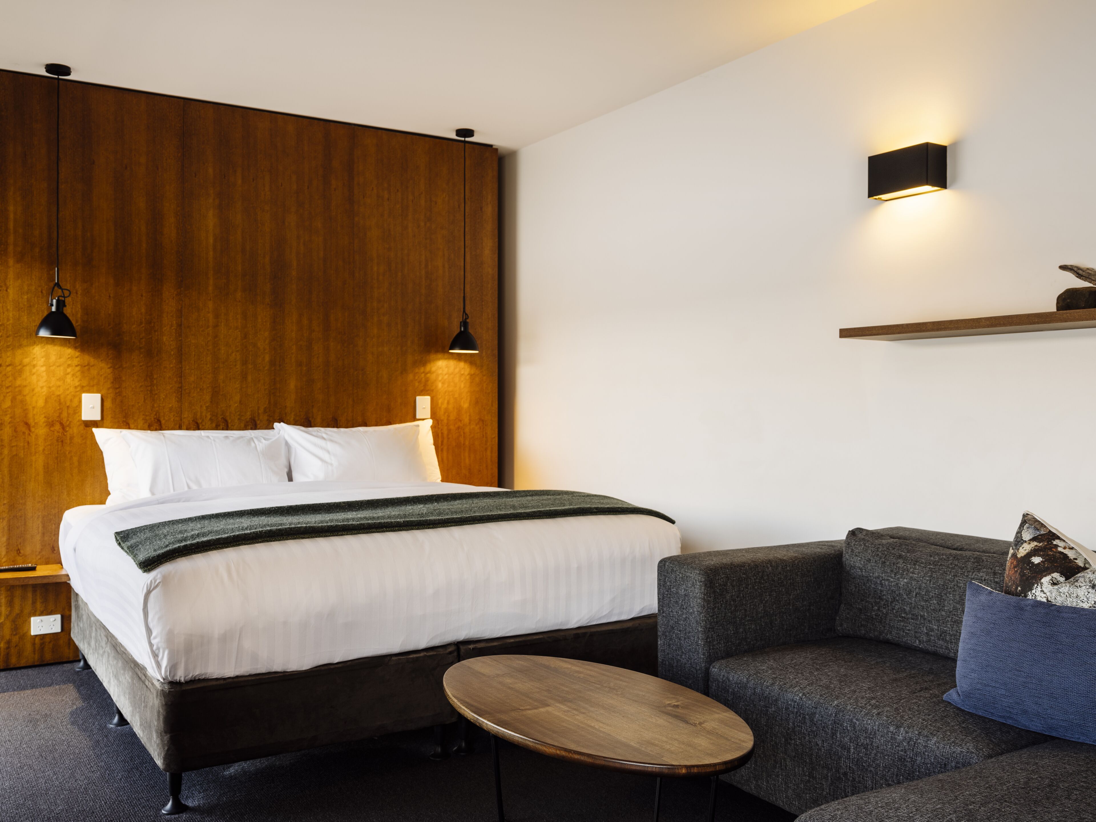 Una camera d'albergo moderna con una testiera in legno, un letto con lenzuola bianche, un tavolino rotondo e un divano grigio con un cuscino blu.