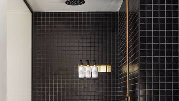 A modern bathroom with black tiled walls, a showerhead, and three bottles of shampoo on a shelf.