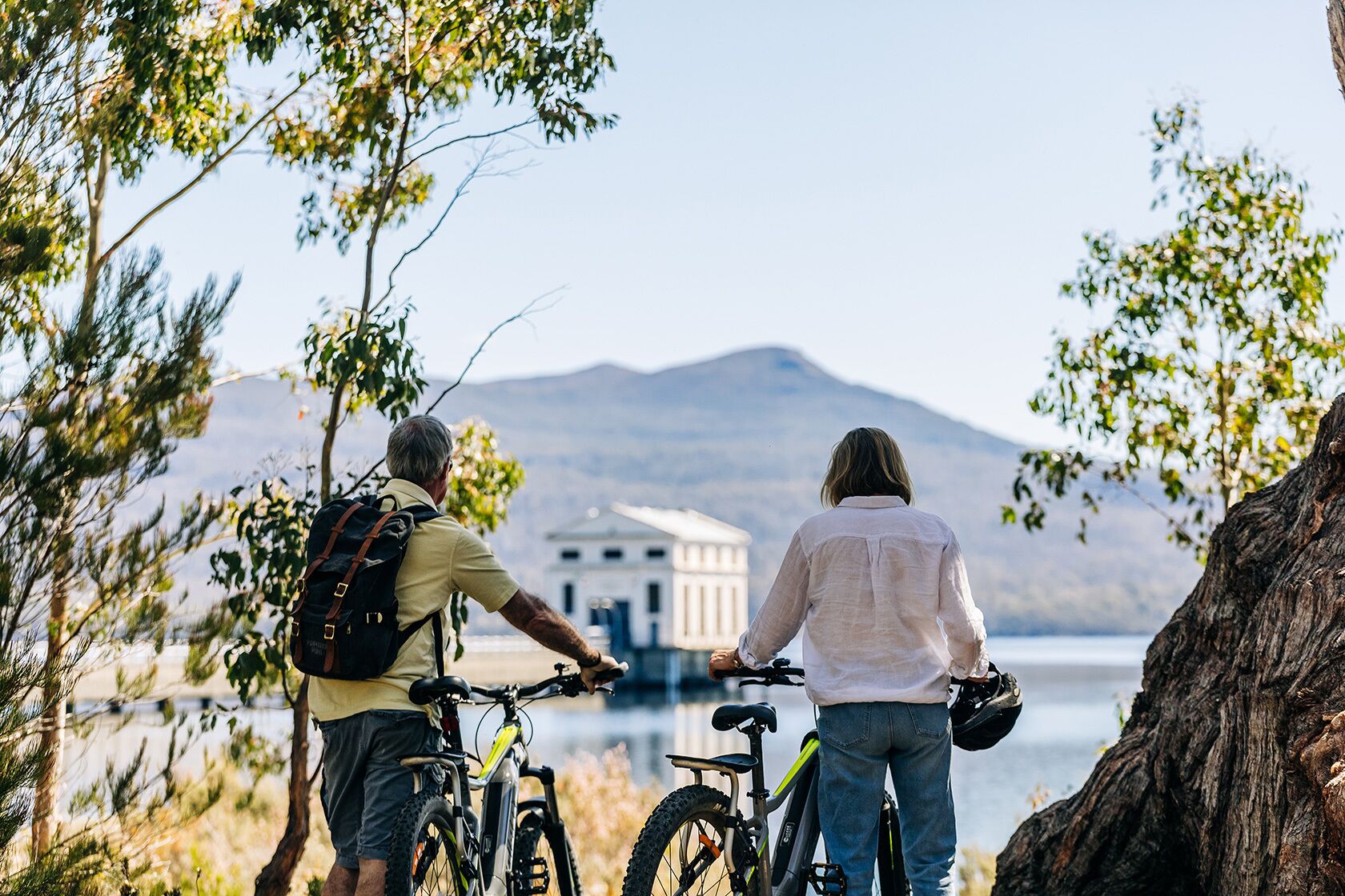 Two people with bicycles, a large tree, and a building with columns by a body of water.