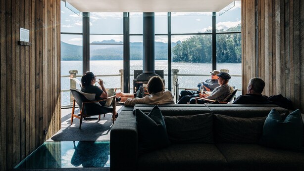 Un interior moderno con amplios ventanales que dan a un lago, paredes de madera y una zona de estar con una mesa de cristal.