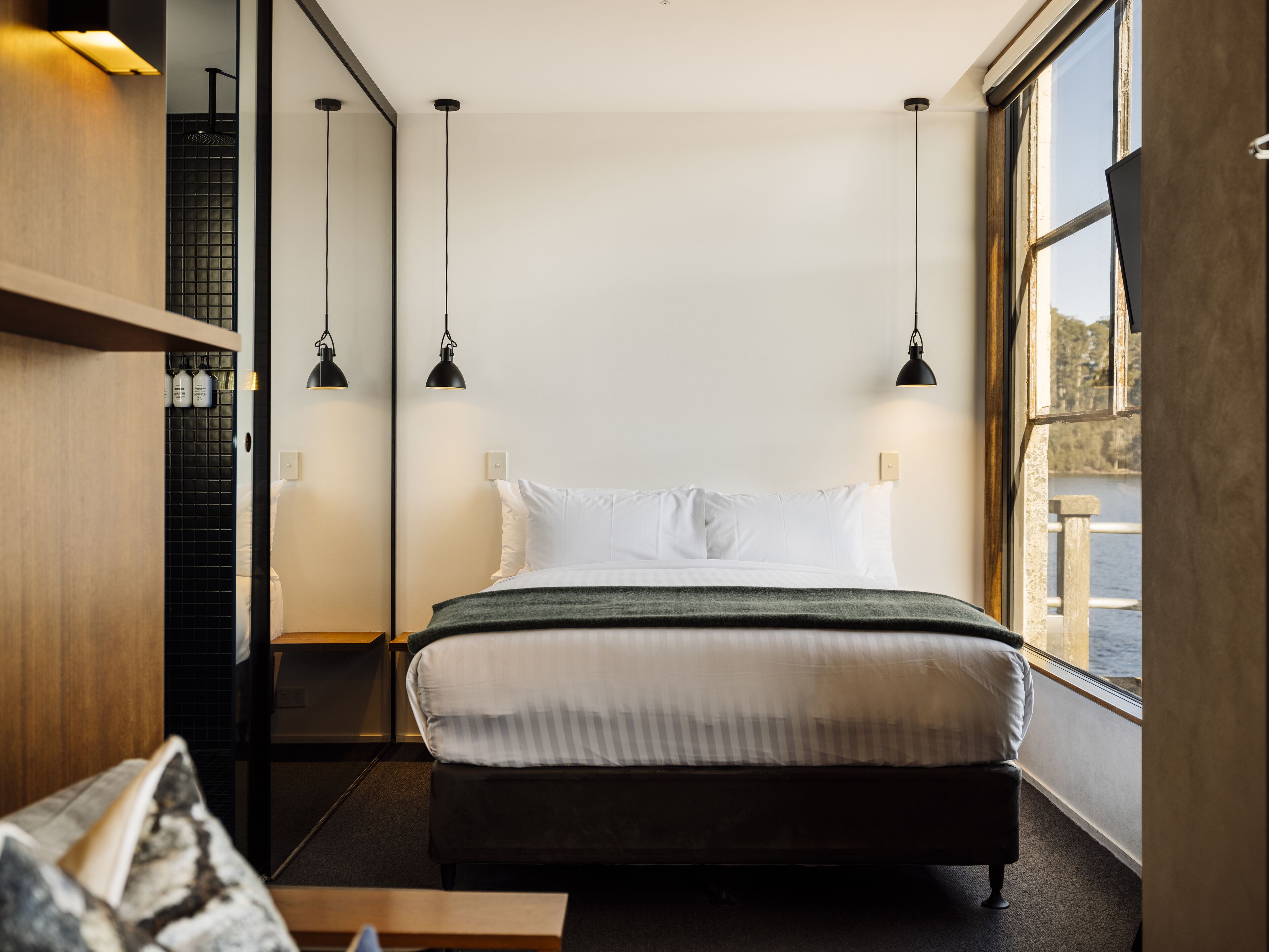 A modern hotel room with a large bed, two pendant lights, a view of the water, and a wooden shelf with bottles.