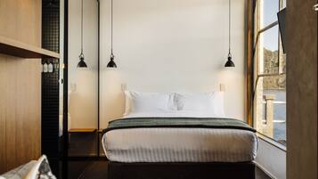 A modern hotel room with a large bed, two pendant lights, a view of the water, and a wooden shelf with bottles.
