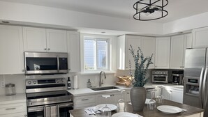 A modern kitchen with white cabinets, stainless steel appliances, a wooden island table, and a large window providing natural light.