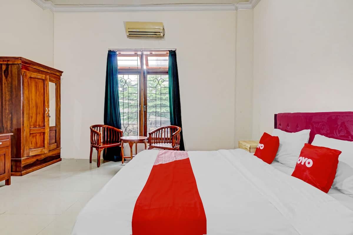 A hotel room with a bed, a wooden wardrobe, two chairs, a small table, and a window with curtains.