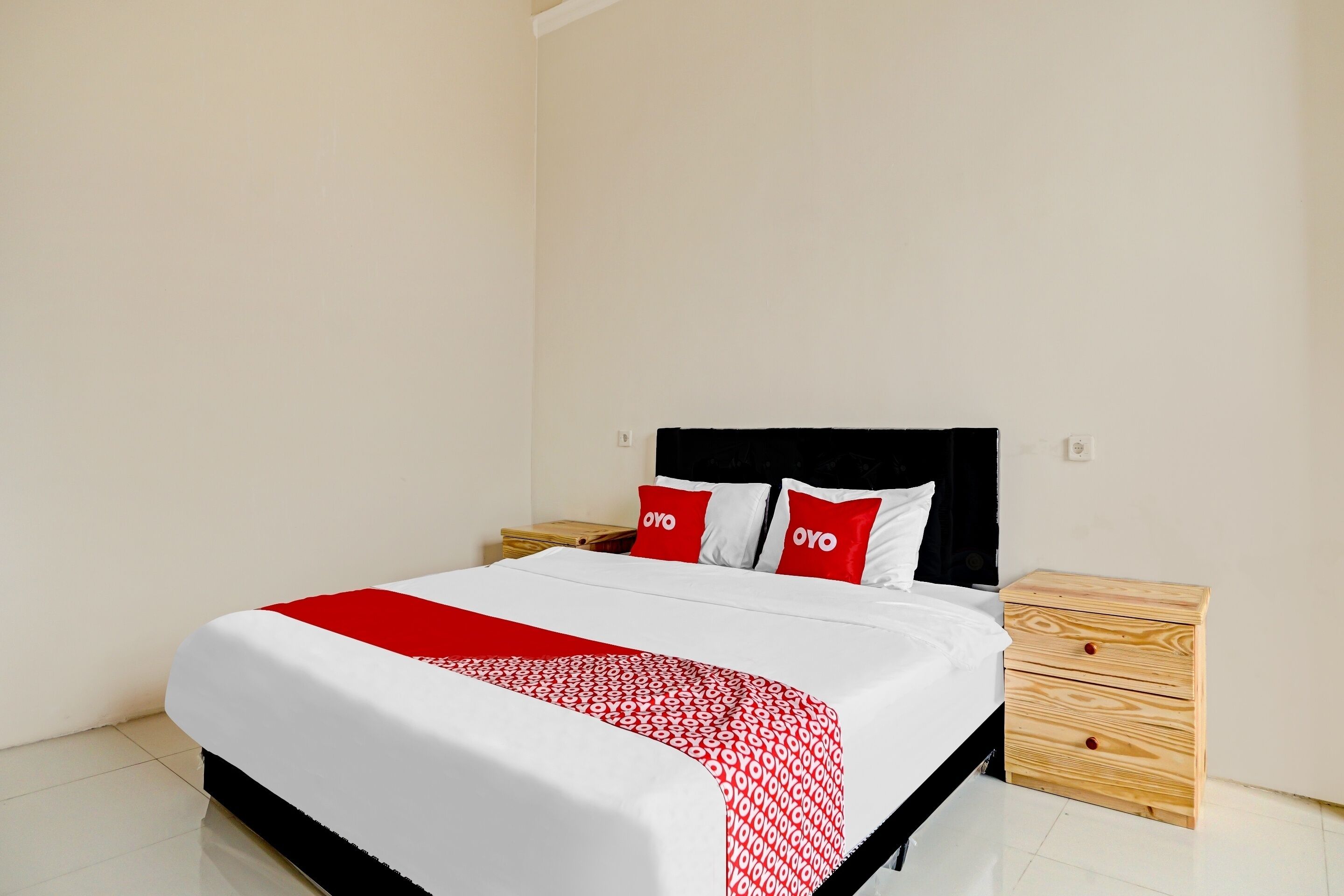 Un letto rifatto con cura, con copriletto bianco e rosso, due cuscini rossi con il marchio "OYO" e un comodino in legno con cassetti.