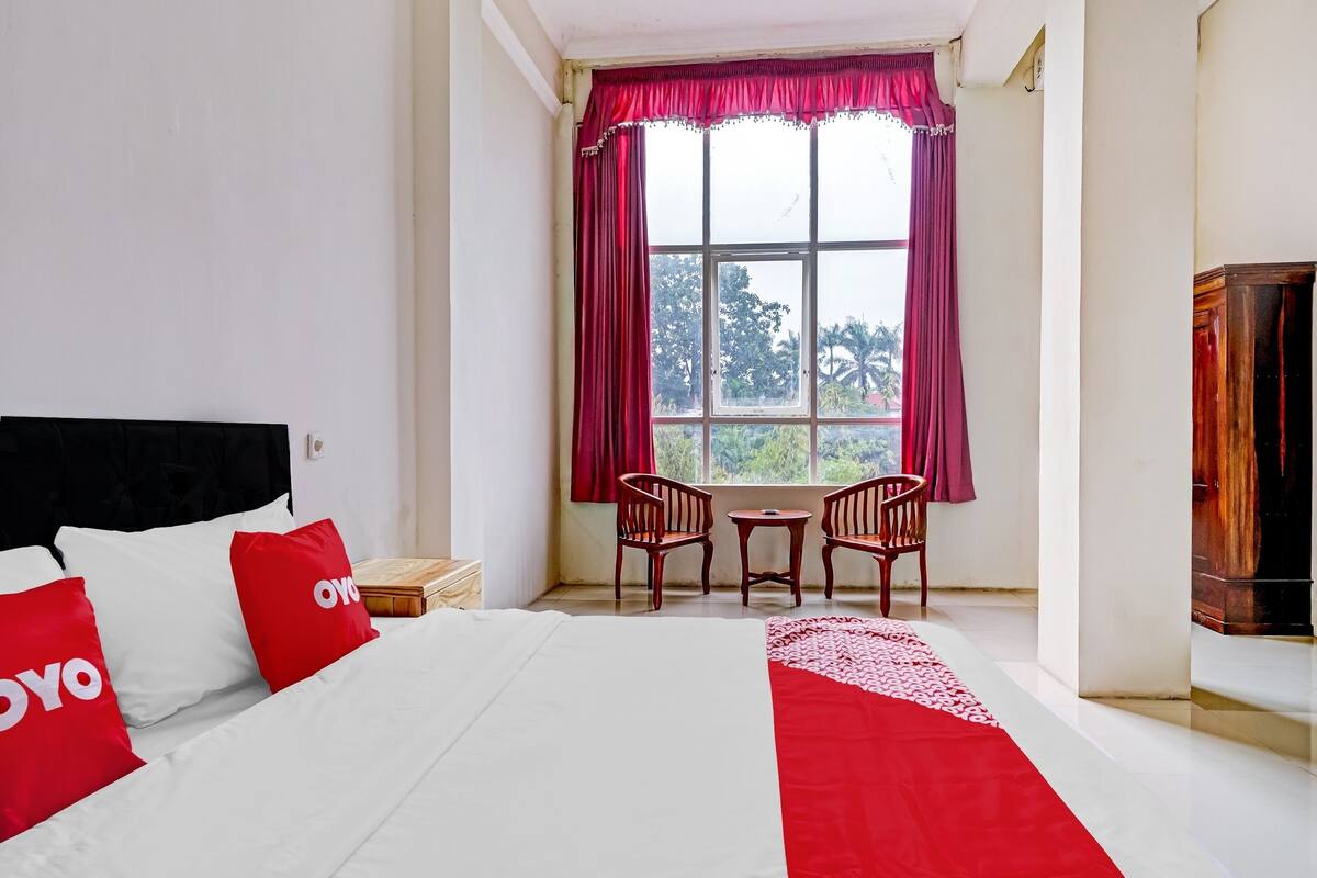 A hotel room with a bed, two red pillows, a window with a view, a small table with chairs, and a wooden cabinet.