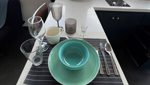 A dining table set with a teal bowl, plates, glasses, and cutlery. A small black speaker and a tissue roll are on the table. - Oasis Loft 5min from SJO airport cozy and comfy fully equipment  down town A/C (Alajuela)
