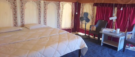 A hotel room with a large bed, a desk with a chair, a fan, and a red curtain.