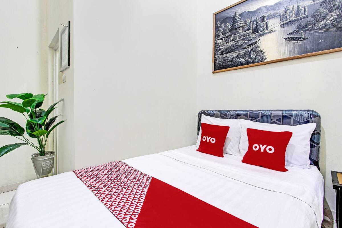 Ein ordentlich bezogenes Bett mit einer roten und weißen Bettdecke, zwei rote Kissen mit dem Aufdruck "OYO" und ein gerahmtes Landschaftsbild an der Wand.