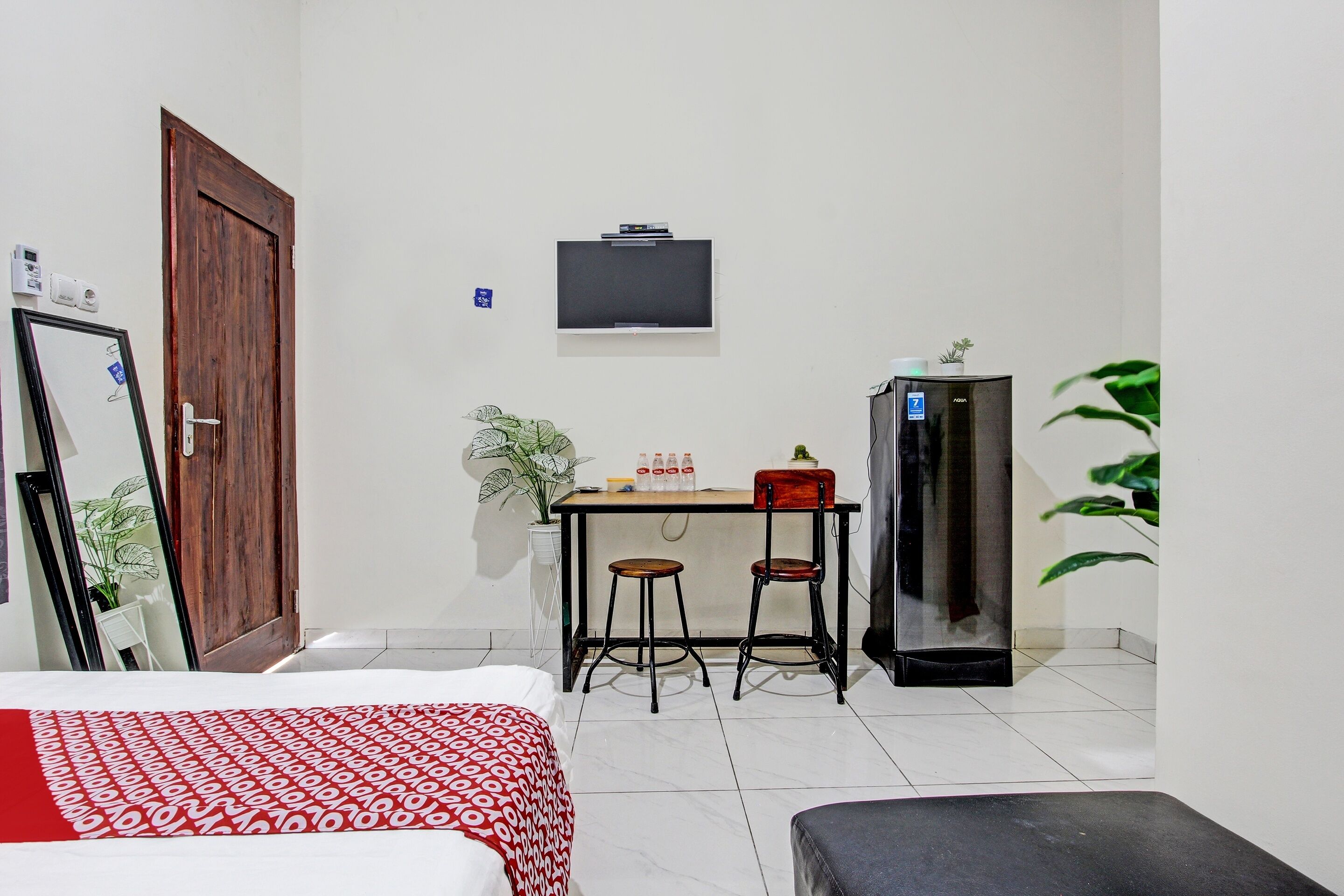 Ein kompaktes Hotelzimmer mit Bett, Schreibtisch mit zwei Stühlen, Fernseher, Kühlschrank und einer Topfpflanze.