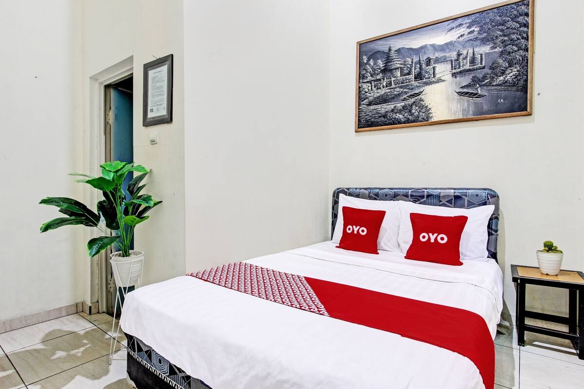 Ein ordentlich eingerichtetes Hotelzimmer mit einem Bett, einer großen Kübelpflanze, einem gerahmten Bild an der Wand und einem kleinen Tisch mit einer Pflanze.