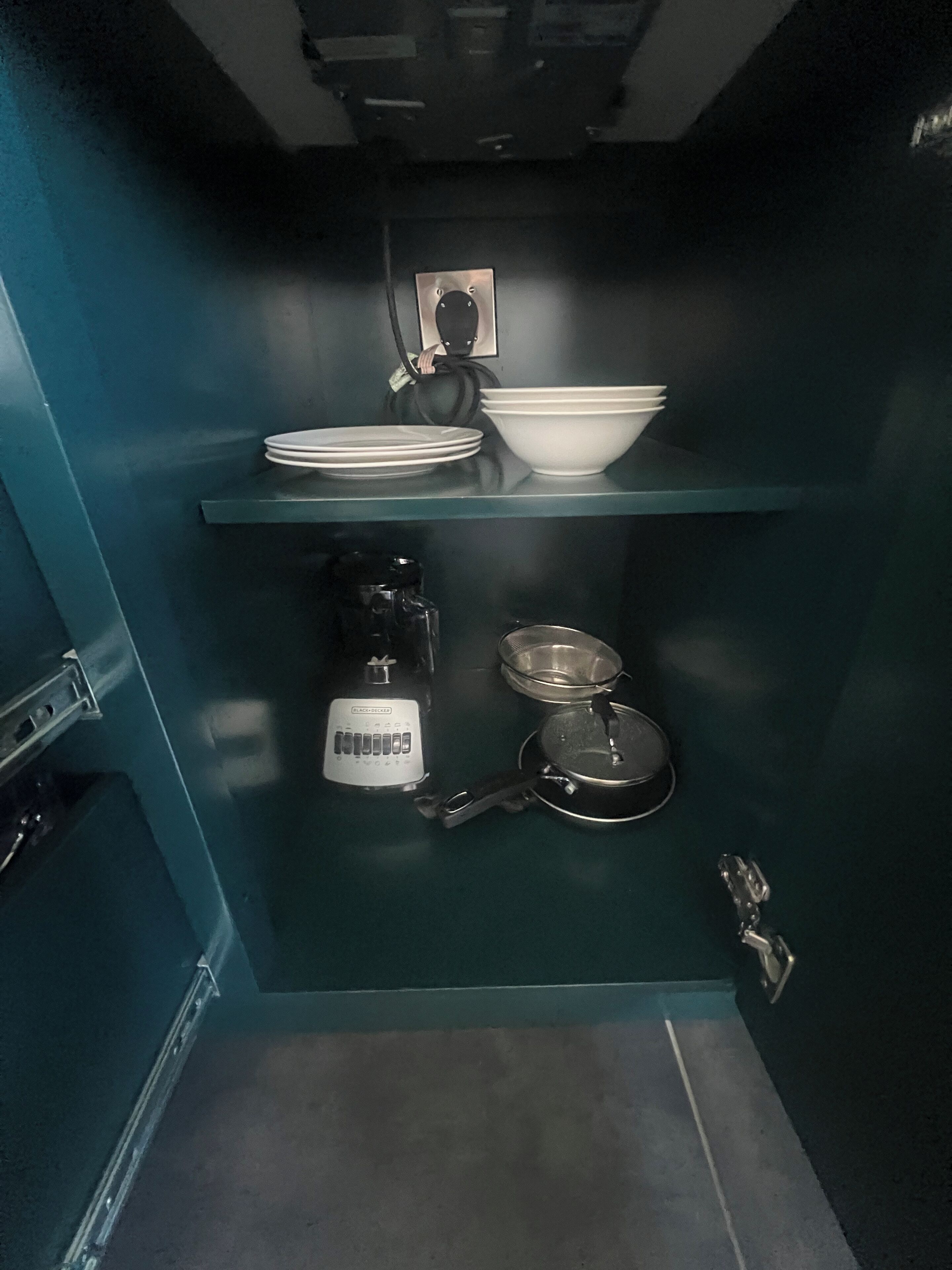 A compact kitchen with a blue wall, a coffee maker, a kettle, and a bowl on a shelf.