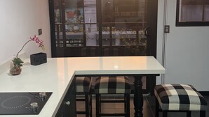 Una cocina con encimera blanca, dos taburetes a cuadros, una mesita y vistas a un atardecer a través de una puerta de cristal.