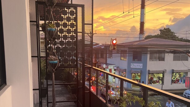 Un balcón con vista a un atardecer, una calle con coches y un edificio con un cartel.