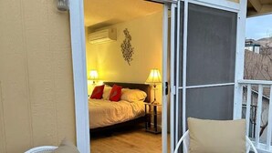 A balcony with two white chairs and a small table, a sliding glass door leading to a bedroom with a bed, two lamps, and a decorative wall piece. - FunStation: 2800sqft Reno Getaway for All Ages (Sparks)