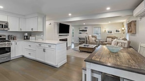 A modern kitchen with white cabinetry, a built-in oven, and a microwave. There is a dining area with a wooden table and chairs, and a living room with a sofa and a TV. - Classic Renovated Cape Cod Getaway (Falmouth)