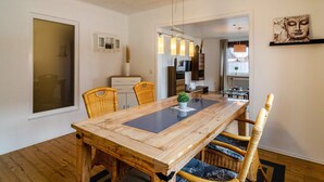 Uma sala de jantar com uma mesa de madeira, cadeiras de vime, um vaso com planta e vista para outro cômodo.