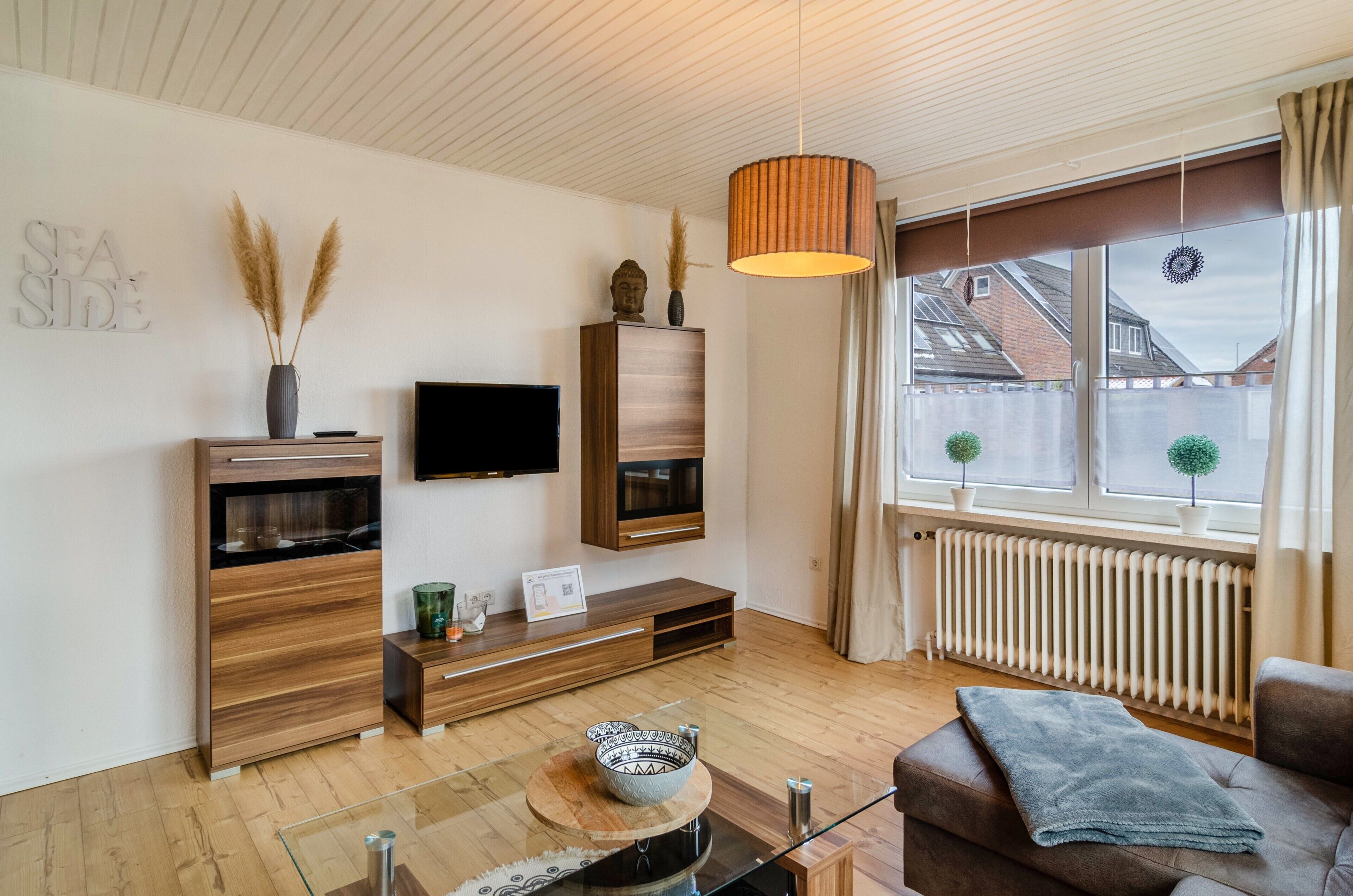 Ein modernes Wohnzimmer mit Flachbildfernseher, Holzmöbeln, einem Glastisch und Blick ins Freie.