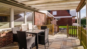 Eine überdachte Terrasse mit Tisch und Stühlen, ein Holzzaun und ein Backsteinhaus im Hintergrund.