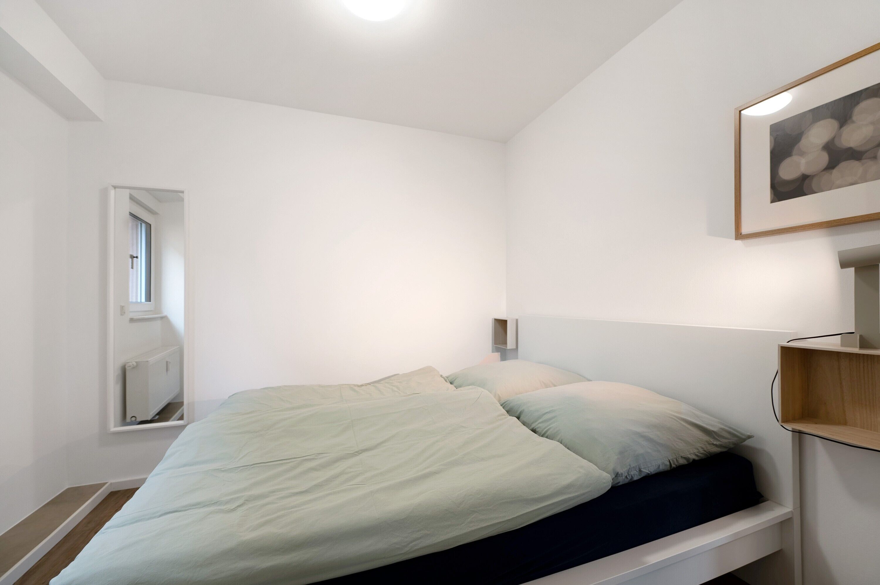 A bedroom with a bed, a window, a radiator, and a framed picture on the wall.
