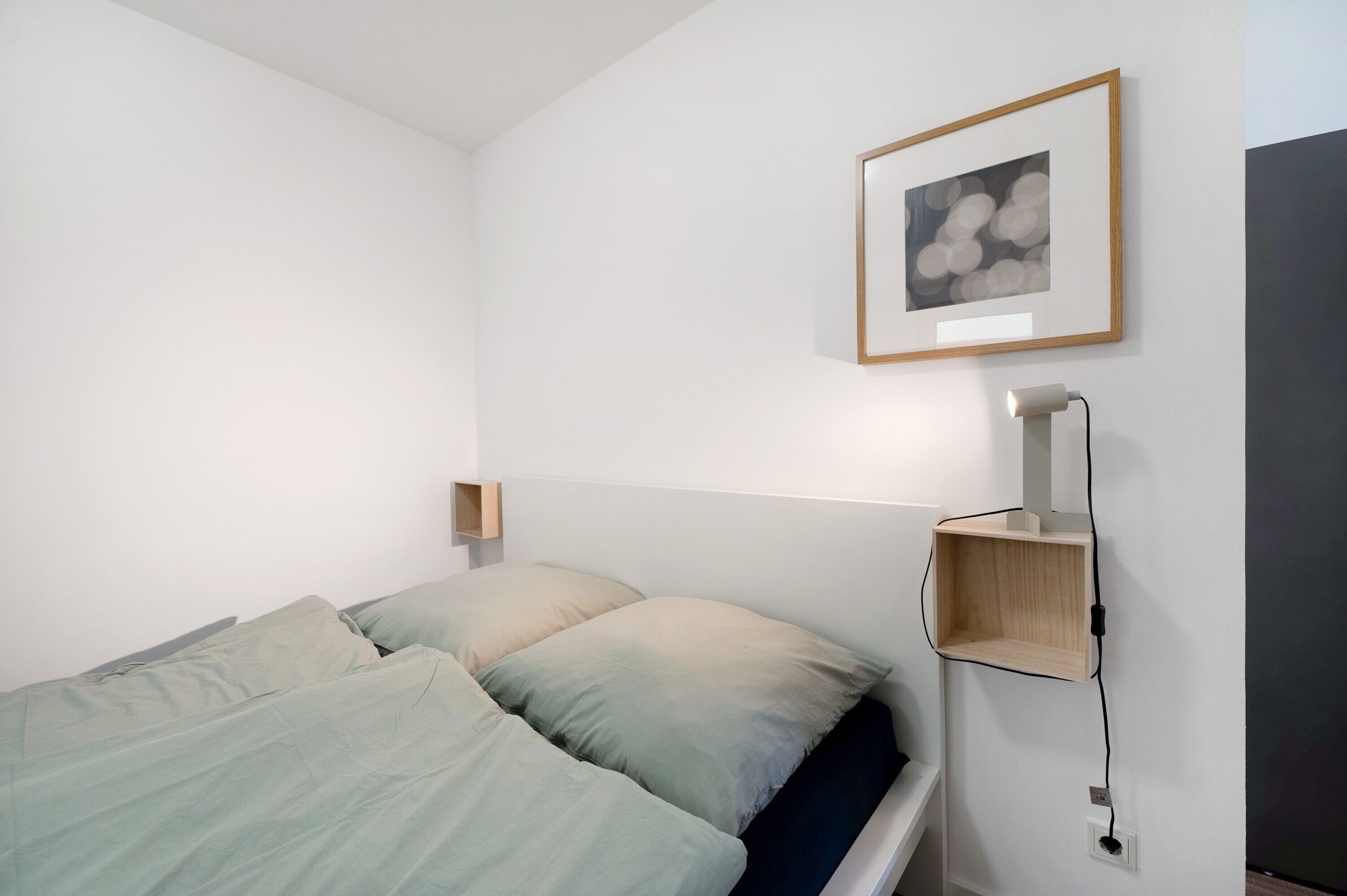 Una cama con ropa de cama verde claro, una mesita de noche con una lámpara y un cuadro en la pared.