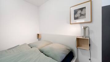 Una cama con ropa de cama verde claro, una mesita de noche con una lámpara y un cuadro en la pared.