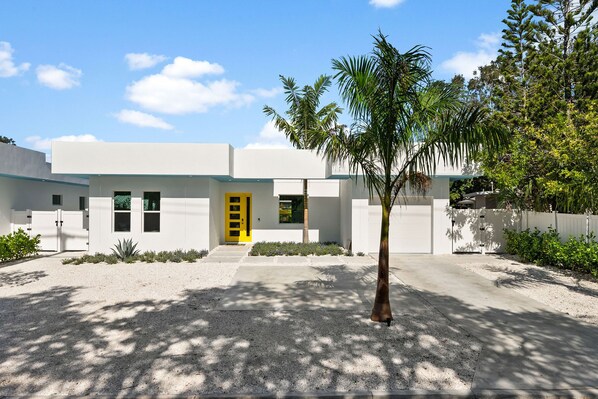 A modern house with a yellow door, surrounded by a gravel driveway and palm trees.