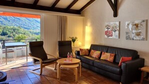 Ein gemütliches Wohnzimmer mit einer schwarzen Ledersofa, zwei Sesseln, einem Holztisch und Blick auf eine bergige Landschaft durch ein großes Fenster.