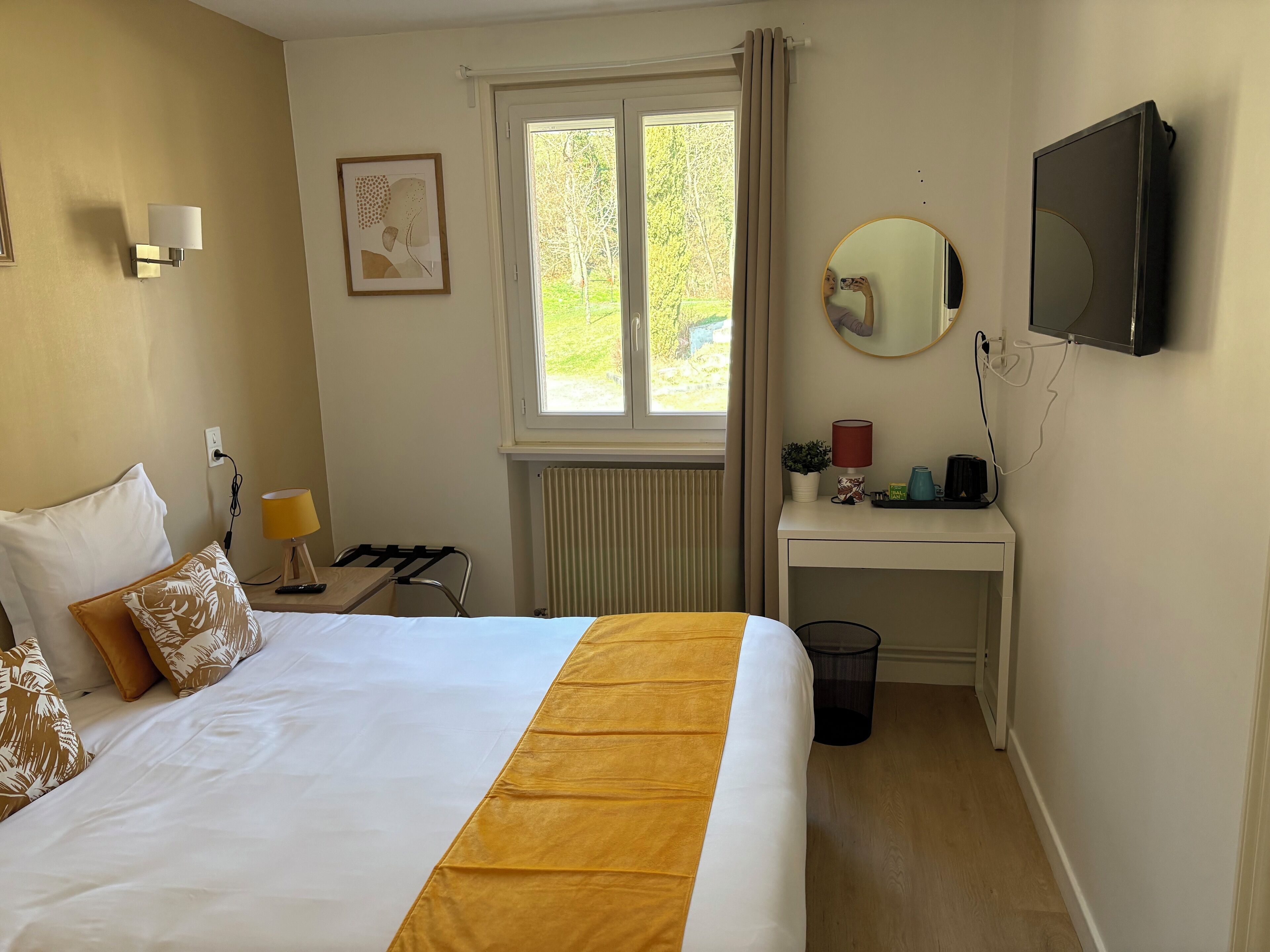 Habitación de hotel con cama, escritorio, silla, ventana, espejo, televisión y radiador.