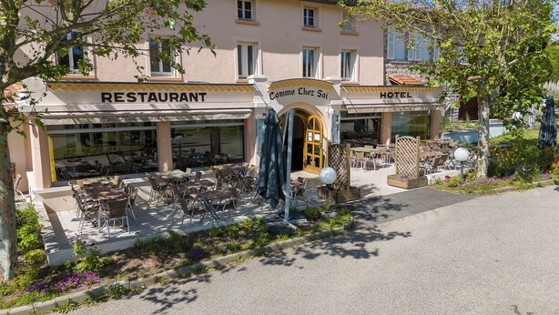 A building with a restaurant and hotel sign, outdoor seating with tables and chairs, and a garden area with flowers. - Comme chez soi (Jas)
