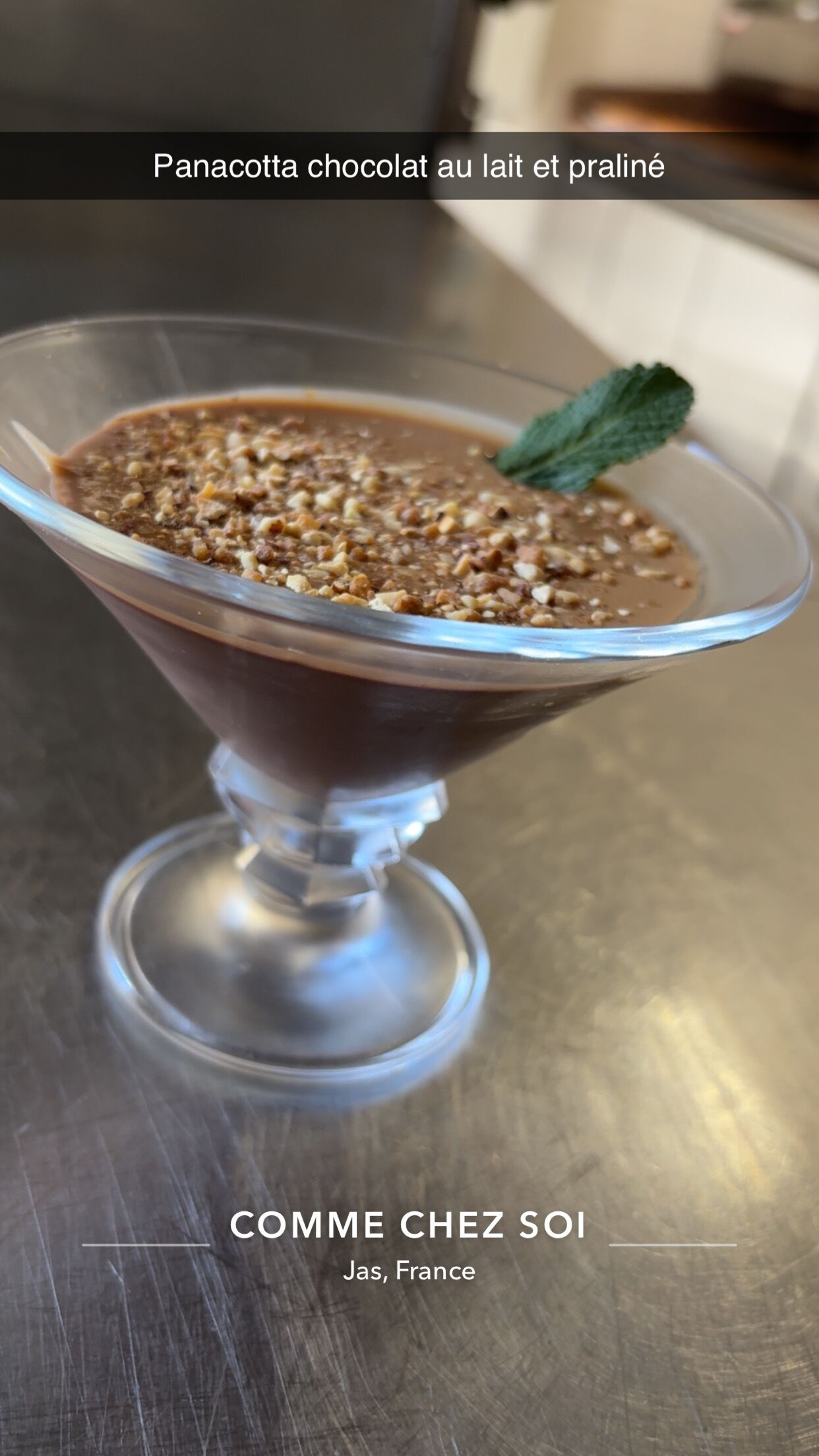 A dessert in a clear glass bowl with a mint garnish, labeled 'Panacotta chocolat au lait et praline' and 'COMME CHEZ SOI Jas, France'.