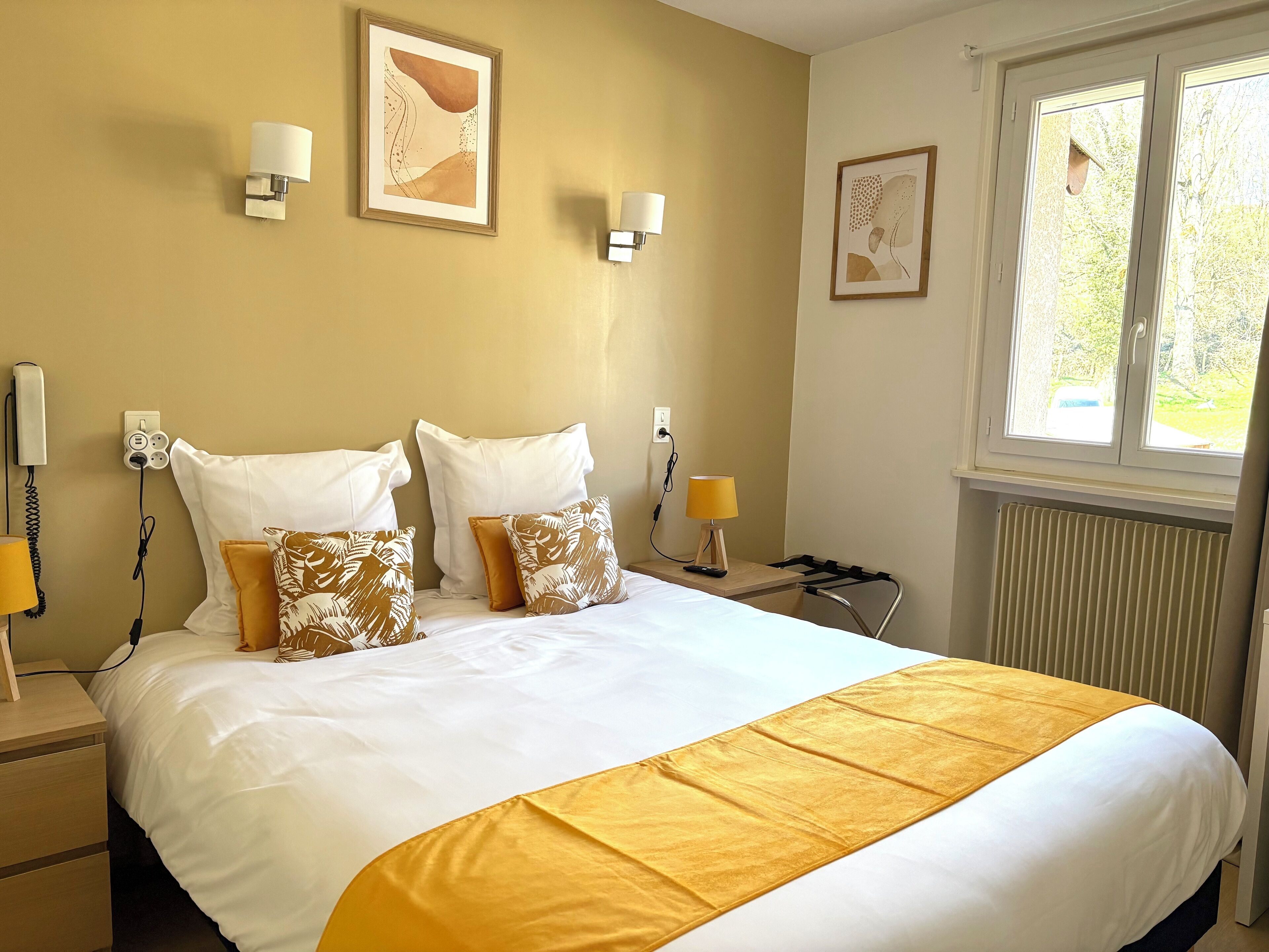 A hotel room with a large bed, two wall-mounted lamps, a radiator, and a window with a view of greenery.