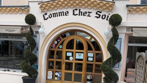 A building entrance with a sign reading "Comme Chez Soir" and a wooden door with glass panels. - Comme chez soi (Jas)